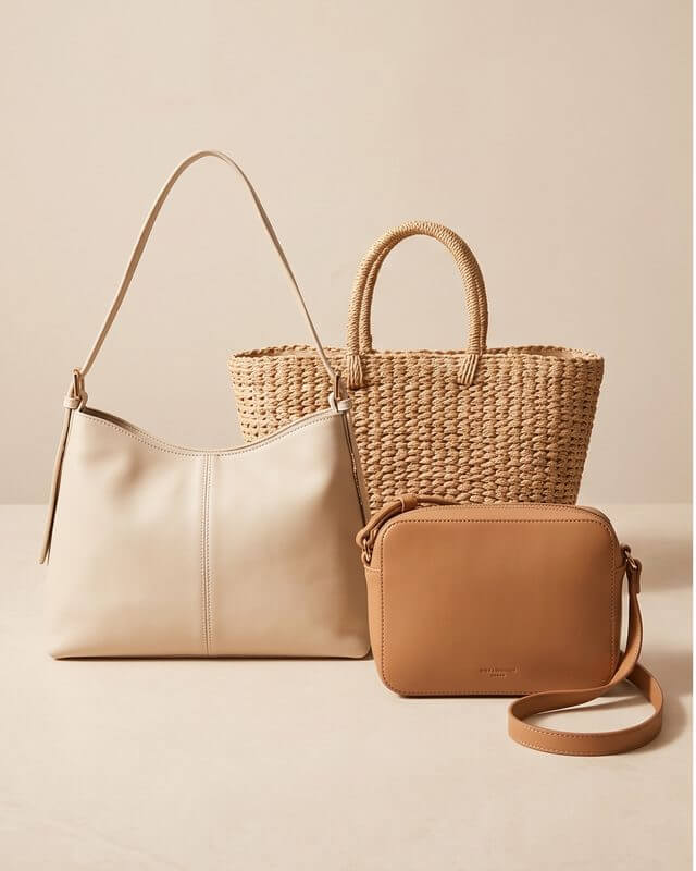 A cream shoulder bag, a woven straw tote and a tan crossbody on a stone surface