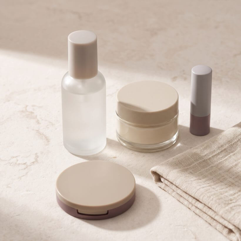 A skincare bottle, cream jar, compact, lip balm and a muslin cloth on stone