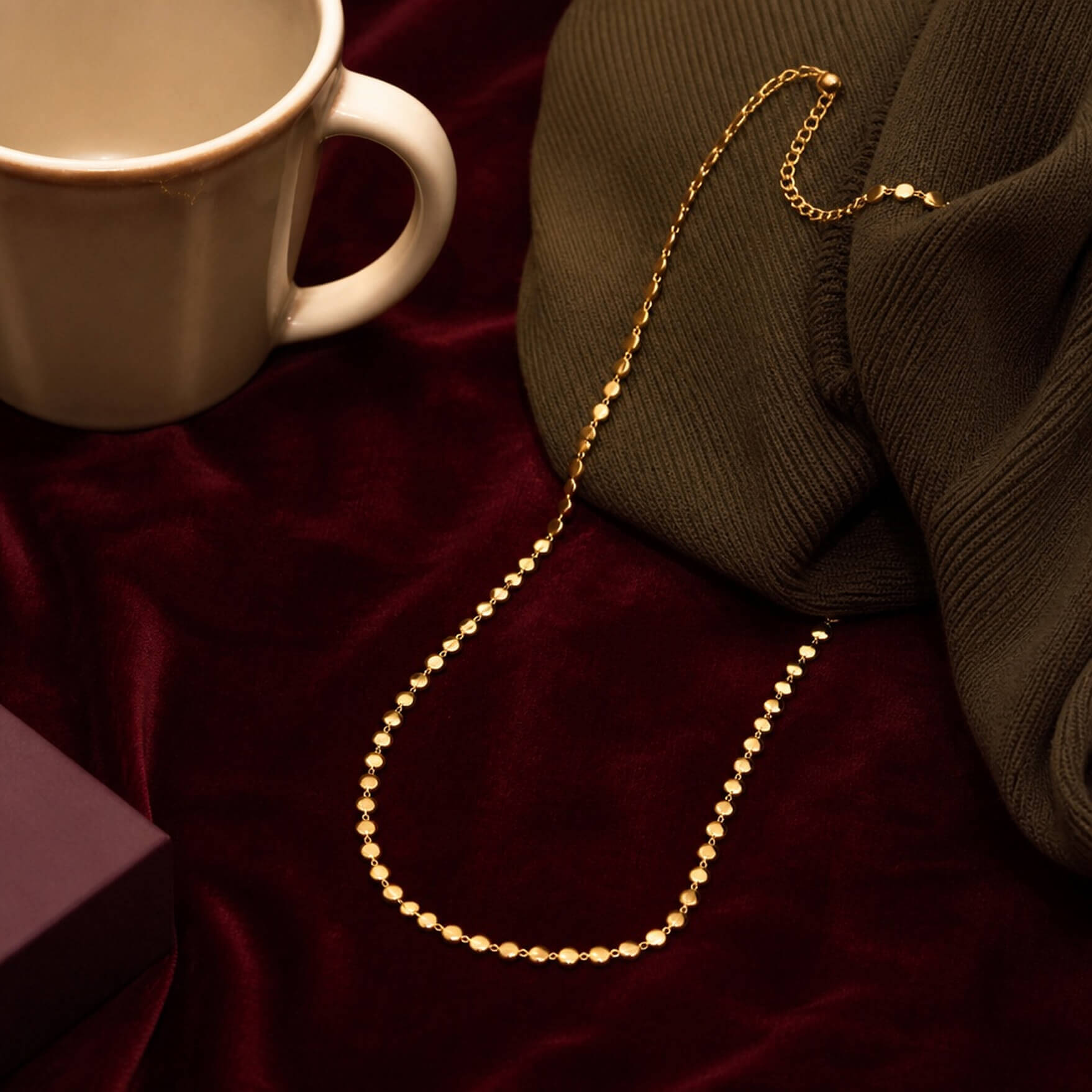 A gold necklace, a folded knit garment and a ceramic mug on a deep burgundy background