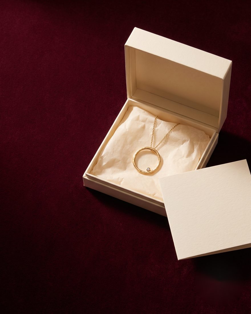 A gold pendant necklace in an open cream gift box on a deep burgundy background