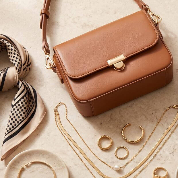 A structured handbag, a printed scarf and layered gold jewelry on a stone surface
