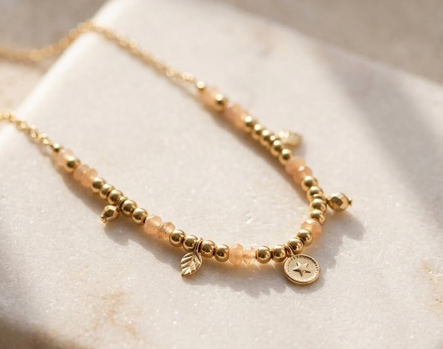 Gold-tone beaded charm necklace with peach stones on a warm stone surface