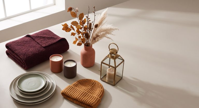 Fall 2026 retail bestsellers on a neutral stone surface: a burgundy boucle throw, stoneware plates, soy candles, a terracotta vase with dried botanicals and pampas, an ochre knit beanie, and a brass lantern.