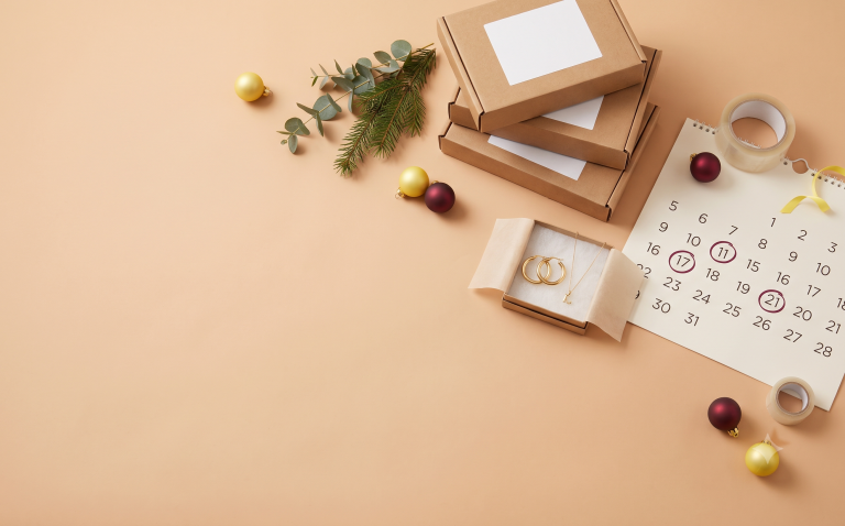 Holiday shipping-prep flat lay — kraft mailer boxes, a wall calendar with key dates circled, and gold jewelry in a gift box among ornaments.