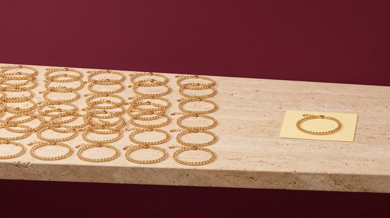 Rows of identical gold curb chain bracelets filling a travertine ledge, with one single bracelet on a pale yellow card set apart, against a deep burgundy wall