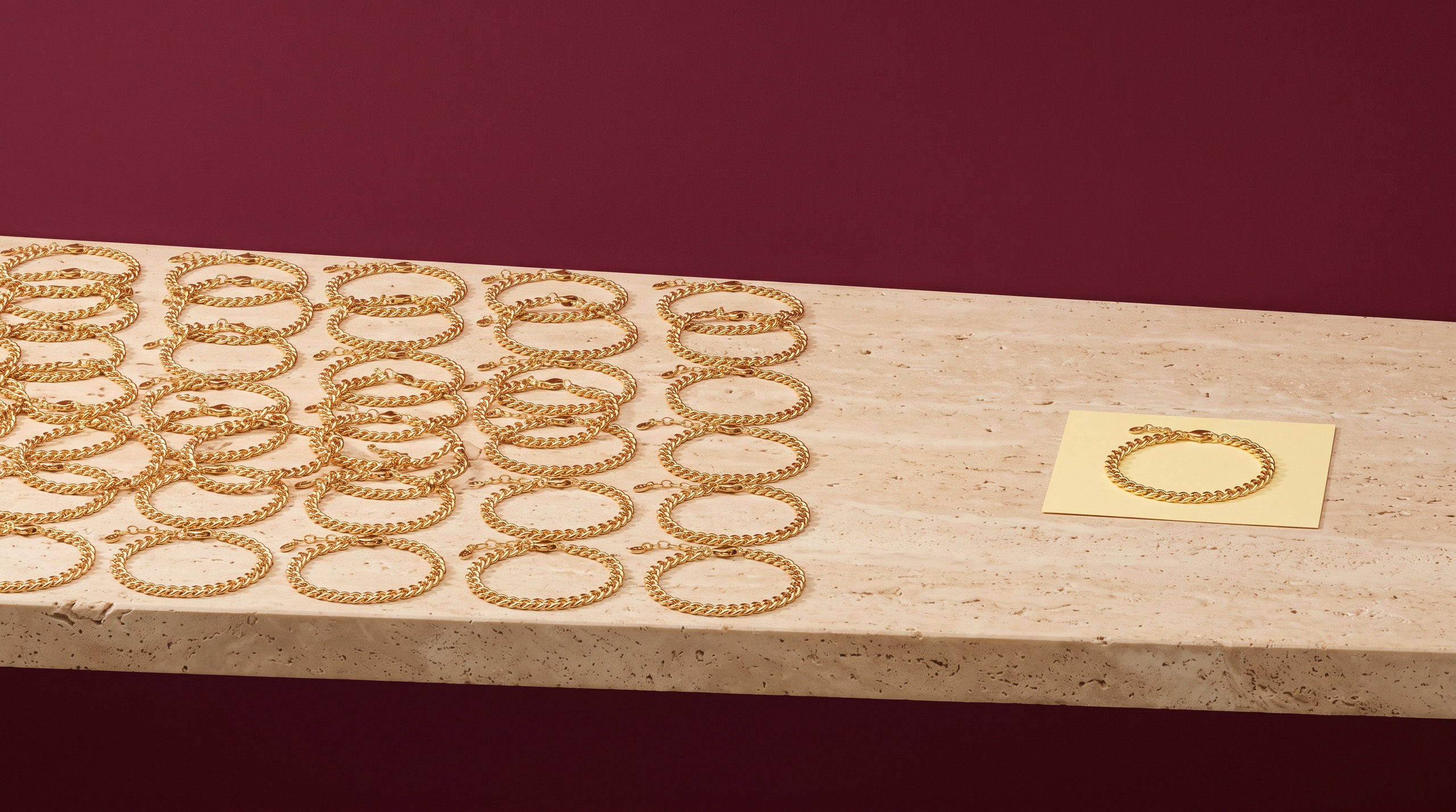 Rows of identical gold curb chain bracelets filling a travertine ledge, with one single bracelet on a pale yellow card set apart, against a deep burgundy wall