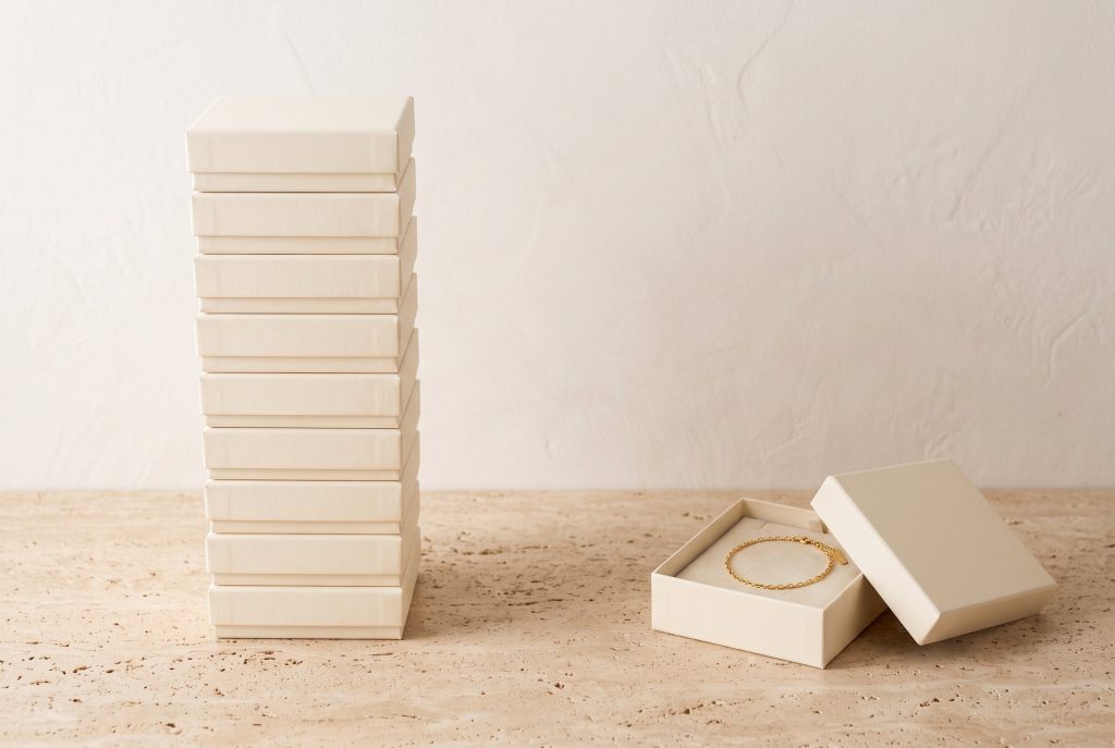 A tall stack of identical cream jewelry boxes beside one open box holding a single gold chain bracelet, on a travertine surface