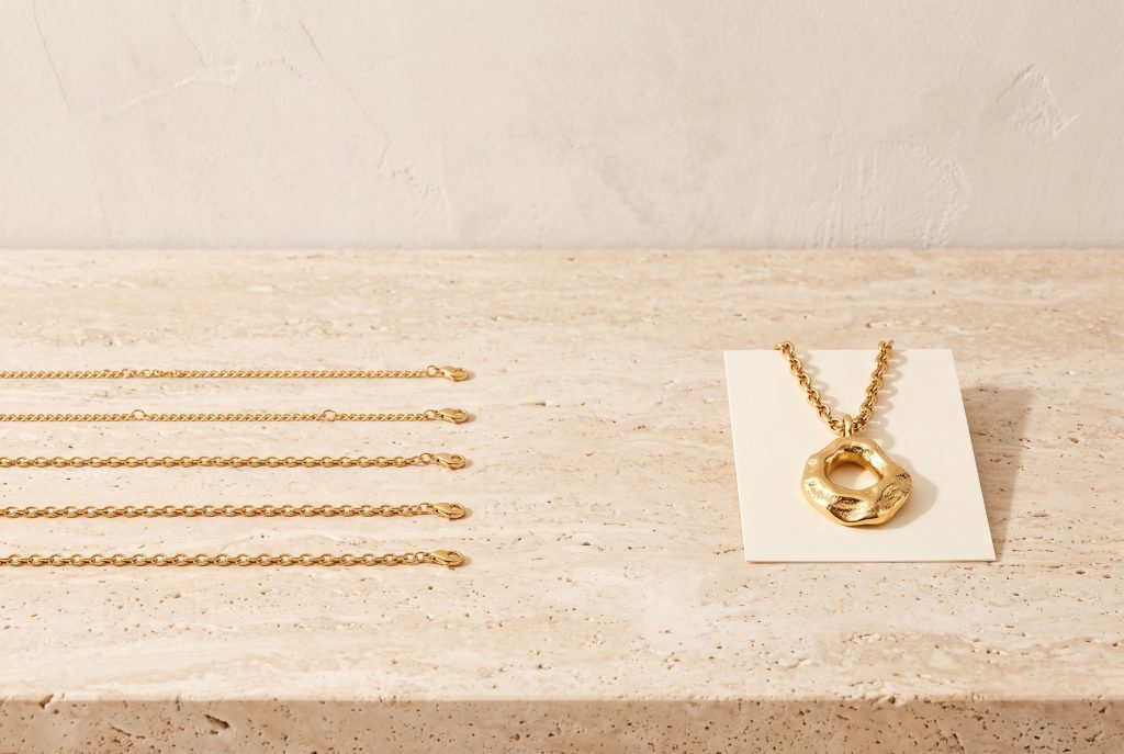 Five identical gold chains laid in rows beside one sculptural textured gold pendant necklace on a blank cream card, on a travertine ledge