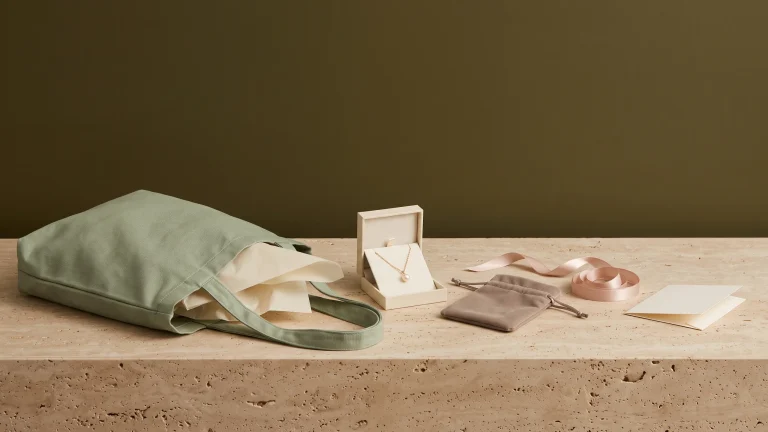 A sage canvas tote lying on its side with cream tissue spilling out, an open gift box holding a gold pearl pendant, a velvet pouch and coiled blush ribbon