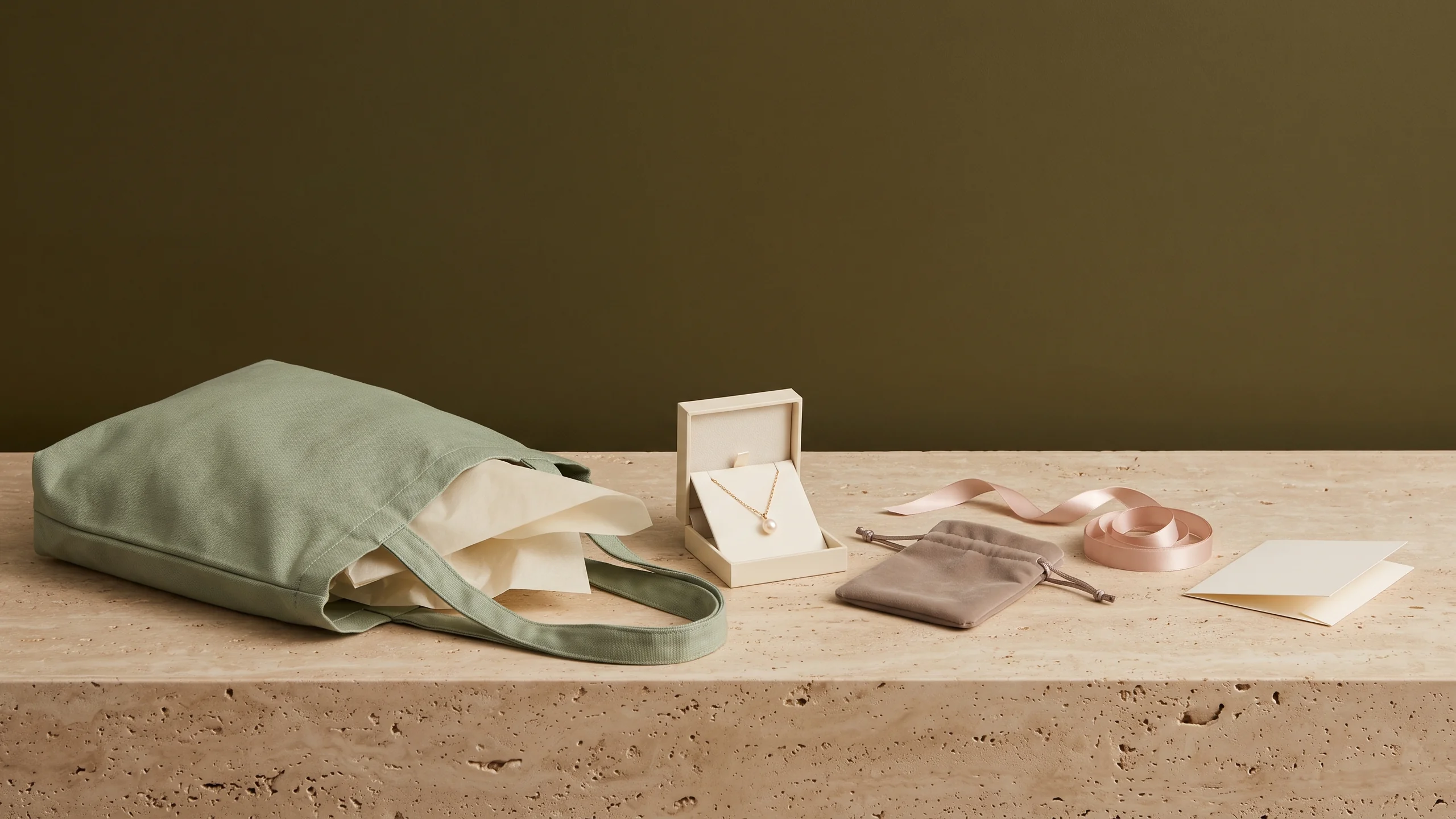 A sage canvas tote lying on its side with cream tissue spilling out, an open gift box holding a gold pearl pendant, a velvet pouch and coiled blush ribbon