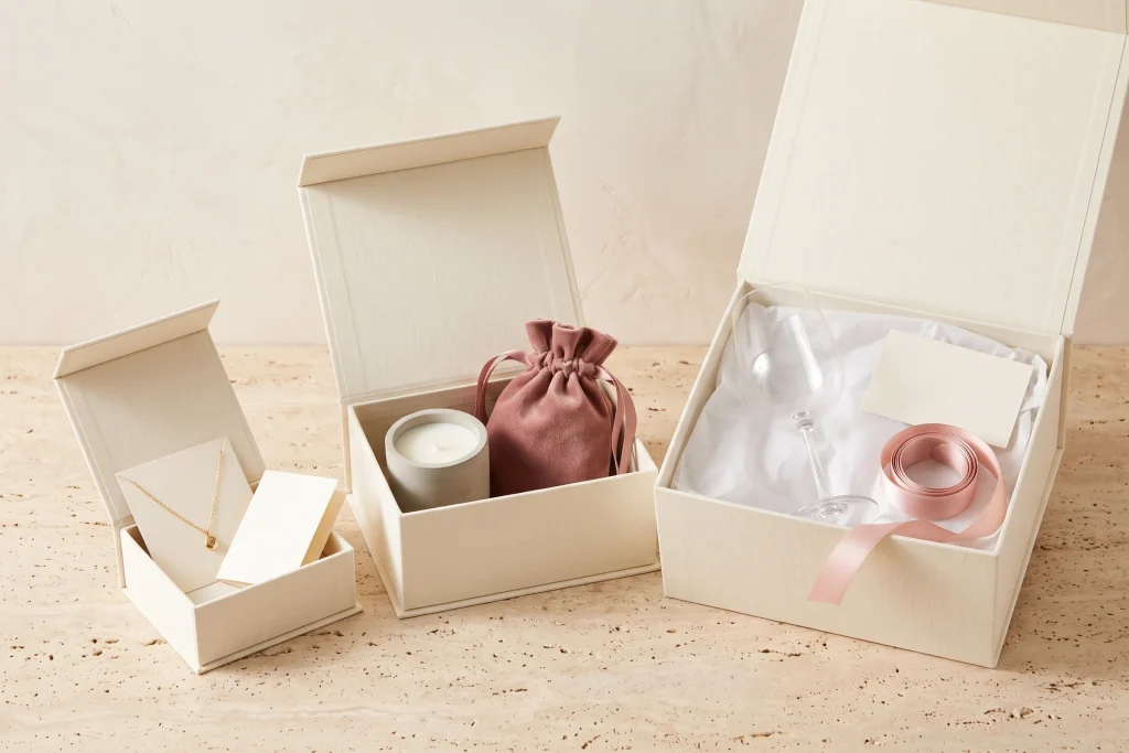 Three open cream gift boxes at three sizes, holding a pendant necklace and card, a candle with a rose velvet pouch, and a stemmed glass with ribbon