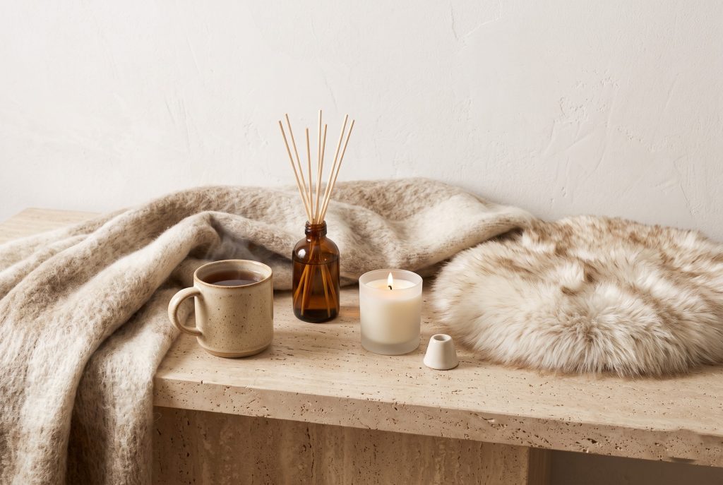 A mohair throw, a faux-fur accent, a reed diffuser, a mug, and a candle in warm neutrals on a stone surface