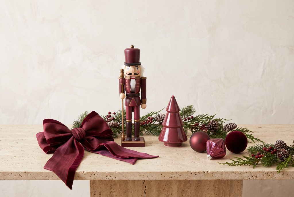 A nutcracker, garland, ceramic tree, ornaments, and a wine-red bow on a stone surface