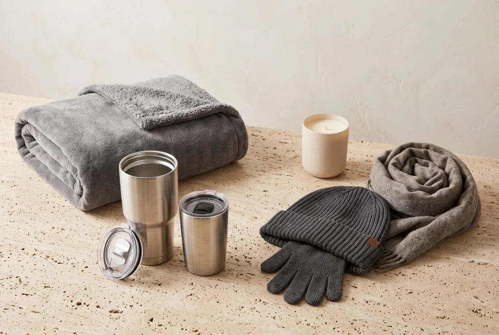A folded grey fleece throw, brushed steel tumbler and travel mug, charcoal knit beanie, gloves and scarf, and a cream candle on travertine