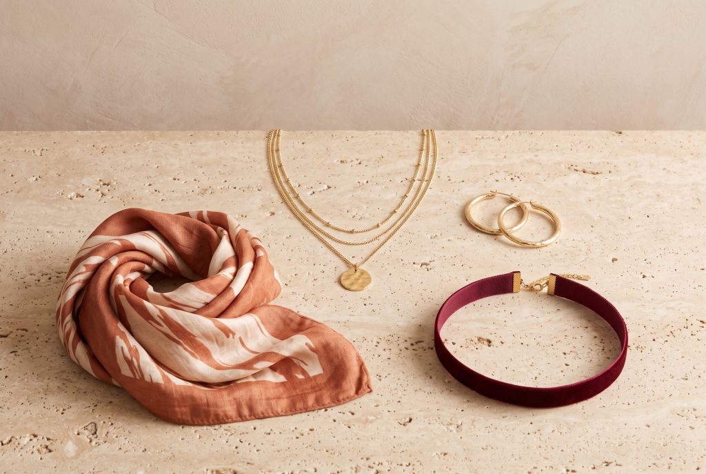 A gold-tone pendant on layered chains, hoop earrings, a velvet choker, and a scarf on a stone surface