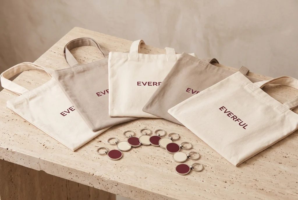 Five cream and taupe Everful tote bags fanned flat across a travertine table, with eleven round burgundy and cream keychains arranged in an arc in front