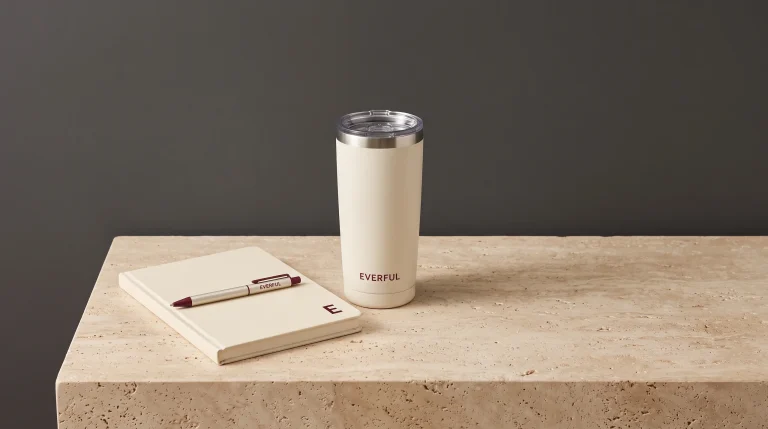 A cream notebook with a small burgundy E on the cover, a branded Everful pen laid across it, and a cream Everful tumbler on travertine against a charcoal wall