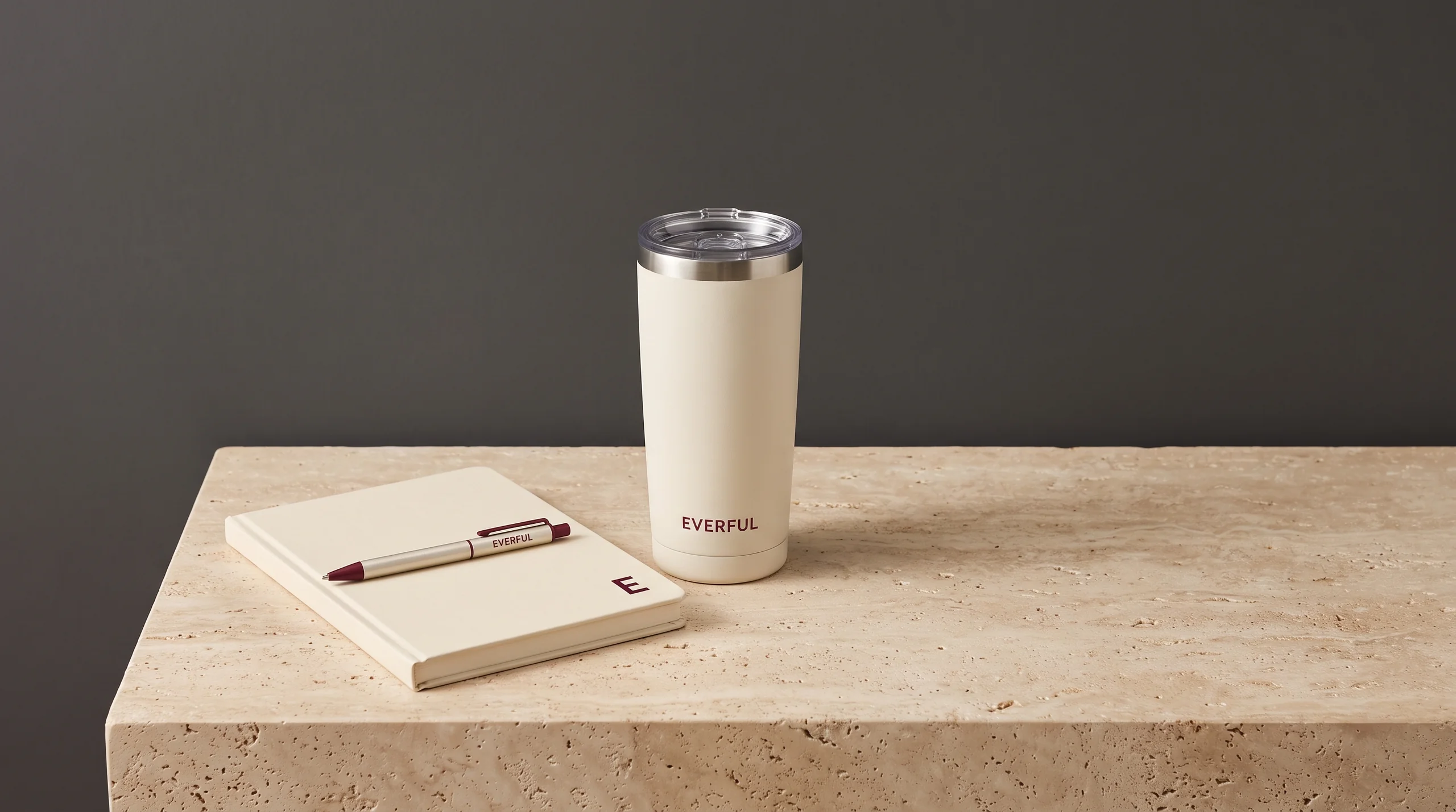 A cream notebook with a small burgundy E on the cover, a branded Everful pen laid across it, and a cream Everful tumbler on travertine against a charcoal wall