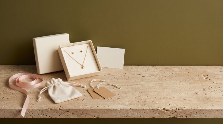 An open cream gift box holding a gold pendant necklace and stud earrings, with a blank card, blush ribbon, and kraft tags on travertine against an olive wall