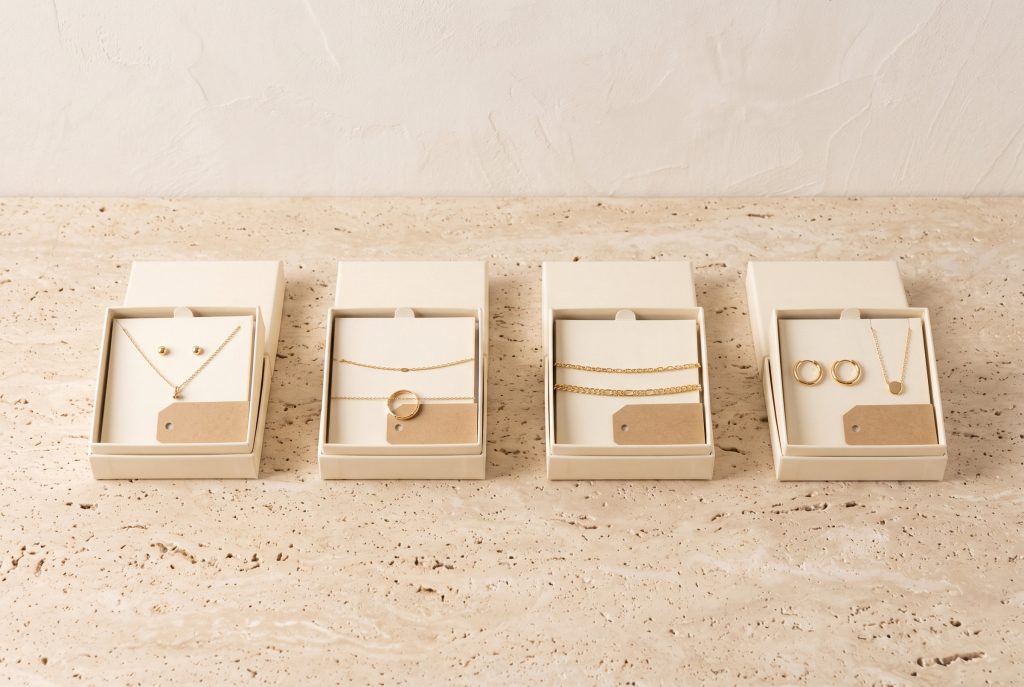 Four open cream gift boxes in a row, each holding a different gold pairing, from studs and a pendant to hoops, chains, and a ring, each with a kraft tag