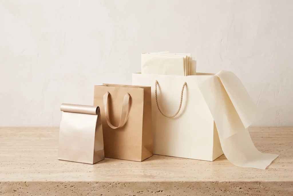 A pearl folded paper bag, a kraft bag with flat handles and a large cream bag with rope handles, holding and draped with cream tissue paper