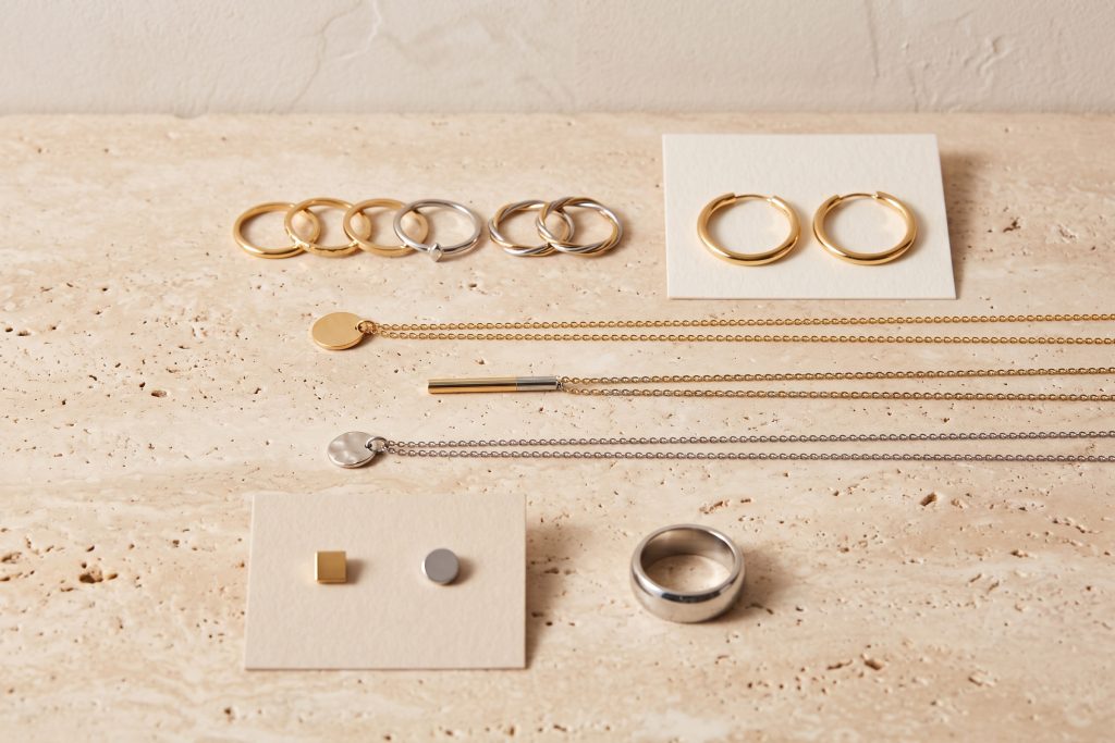 Stacking rings, pendant necklaces, hoop earrings, and studs in gold and stainless finishes arranged in rows on a stone surface