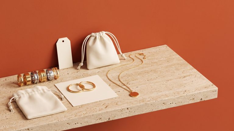 Gold and stainless stacking rings, gold hoops on a blank card, a pendant necklace, and cream pouches on a travertine ledge against a burnt orange wall