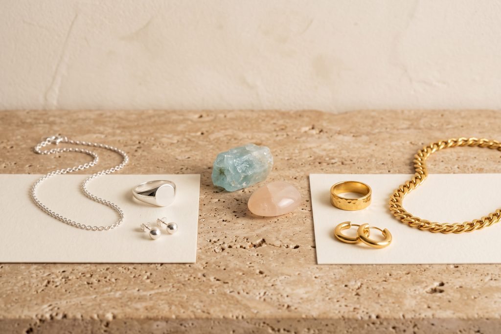 Sterling silver chain, signet ring, and studs on one card beside gold-plated hoops, a textured band, and a curb chain on another, with pale raw stones between them