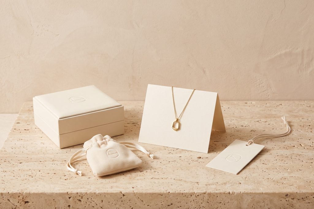 A cream jewelry box, velvet pouch, folded card, and hang tag with a blind-embossed emblem, beside a gold-tone pendant, on a stone surface