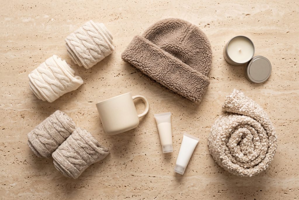 An overhead flatlay of rolled cable-knit socks, a sherpa beanie, a cream ceramic mug, lotion tubes, an open candle tin, and a rolled boucle scarf
