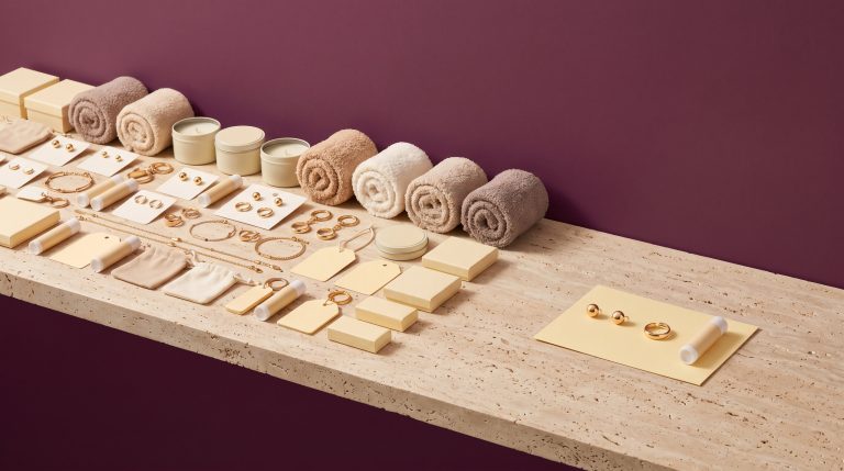 A travertine ledge crowded with gold jewelry, candle tins, lip balms, and rolled socks, a few pieces set apart on a yellow card, against a deep wine wall