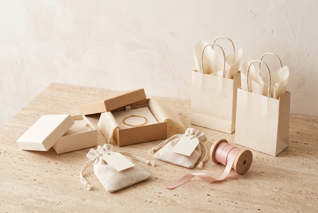 Kraft and cream gift boxes, one holding a gold chain bracelet, with linen drawstring pouches, blush ribbon on a wooden spool, and cream gift bags