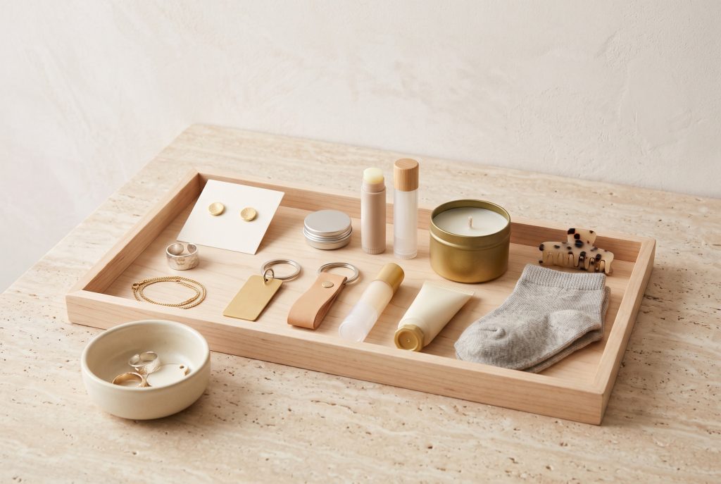 A wooden tray of stud earrings, rings, a gold bracelet, keychains, lip balms, a candle tin, a claw clip, and folded socks, with a ceramic ring dish