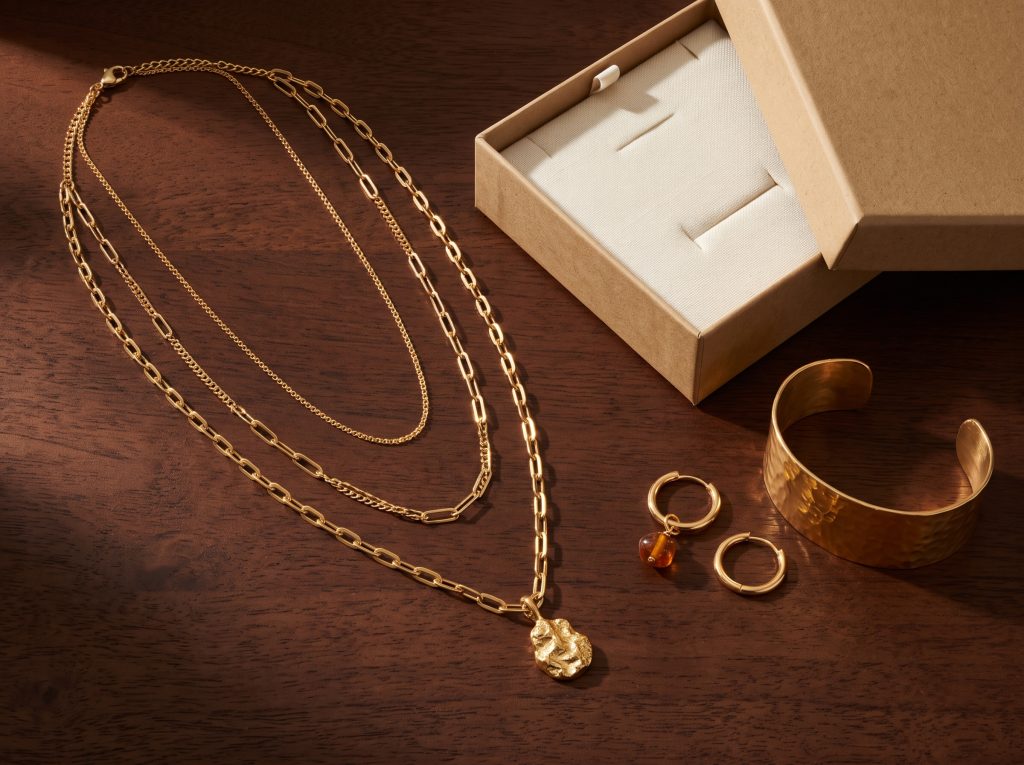 Three gold layering necklaces at graduated lengths beside a kraft gift box on a warm brown surface