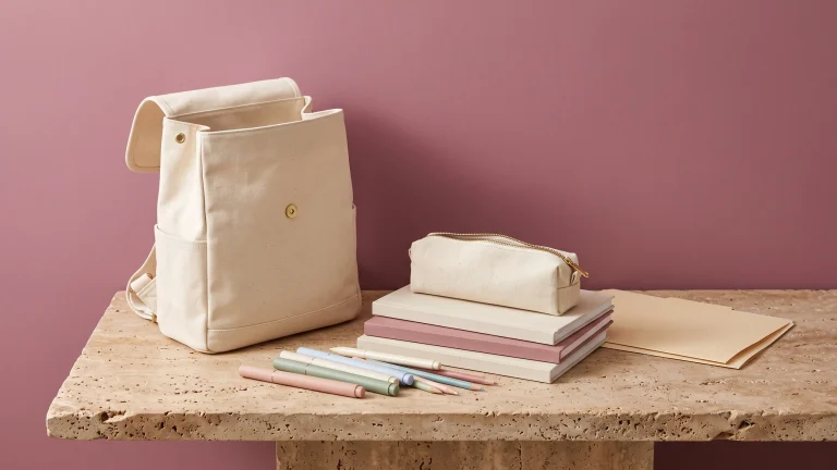 A cream canvas backpack, stacked blank notebooks, a zipped pencil case and loose pastel markers on a stone bench
