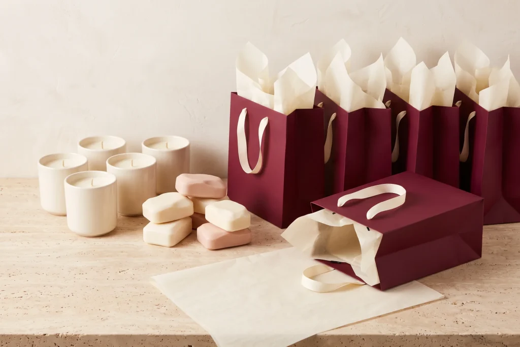 Small cream candles and blush soap bars grouped beside four burgundy gift bags with cream tissue, laid out for assembly on travertine