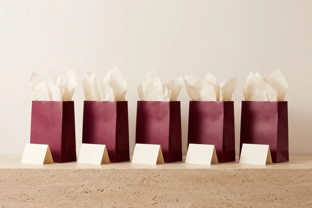 Five matching burgundy gift bags with cream tissue standing in a row, each with a blank cream card propped in front, on travertine