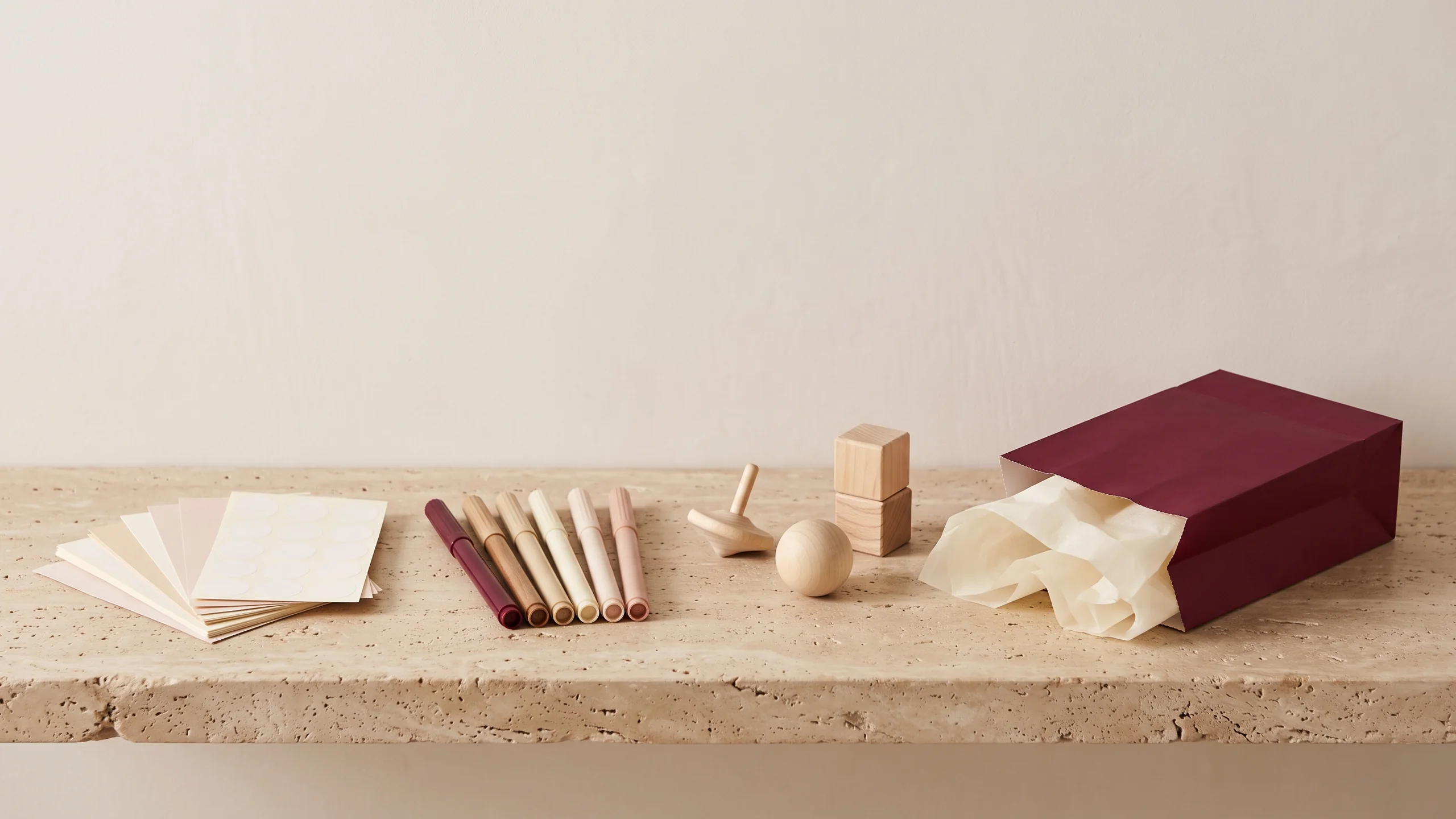 Blank sticker sheets, colored markers and small wooden toys laid out beside an open burgundy goodie bag with cream tissue, on travertine