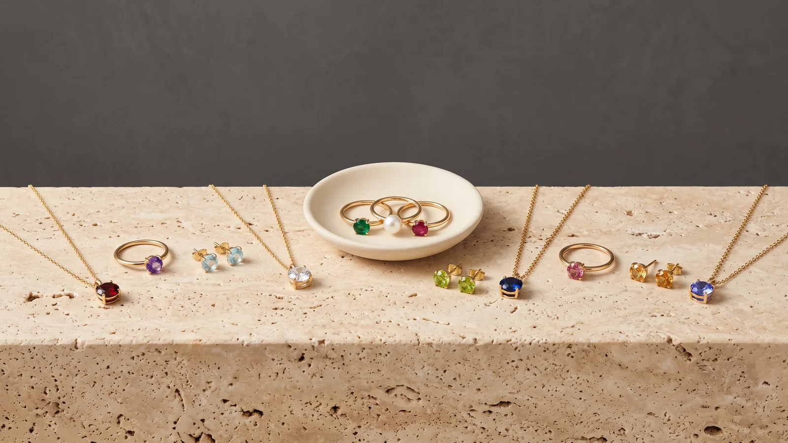 Gold birthstone necklaces, rings and stud earrings in twelve different colored stones on travertine, with three rings on a cream dish