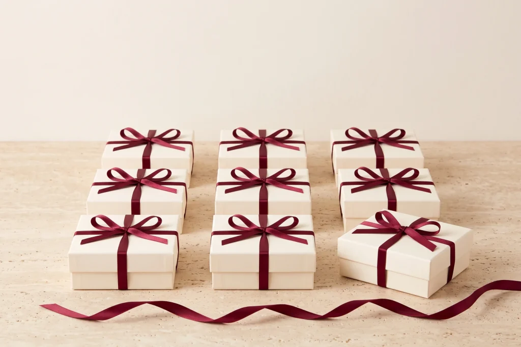 Nine identical ivory favor boxes tied with burgundy satin ribbon, set out in an even grid on travertine with ribbon trailing in front