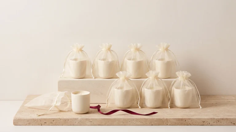 A tiered display of sheer ivory organza bags holding small cream candles, with one empty bag and a loose candle in front, on travertine