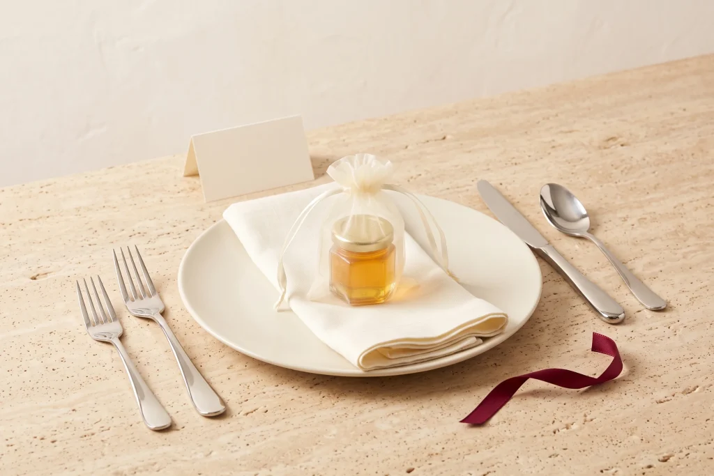 A place setting with a sheer ivory organza bag holding a small jar of honey on a folded napkin, beside a blank cream place card