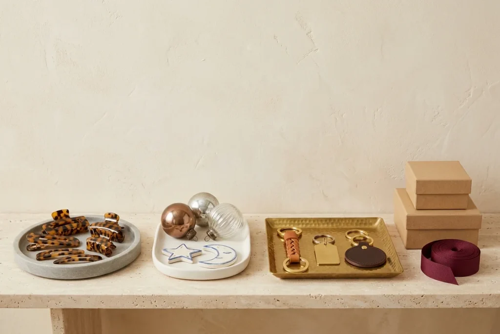 A travertine bench with a stone tray of tortoiseshell hair clips, a white tray of glass ornaments with a ceramic star and moon, a hammered brass tray of leather and brass keychains, stacked kraft gift boxes and a roll of burgundy ribbon