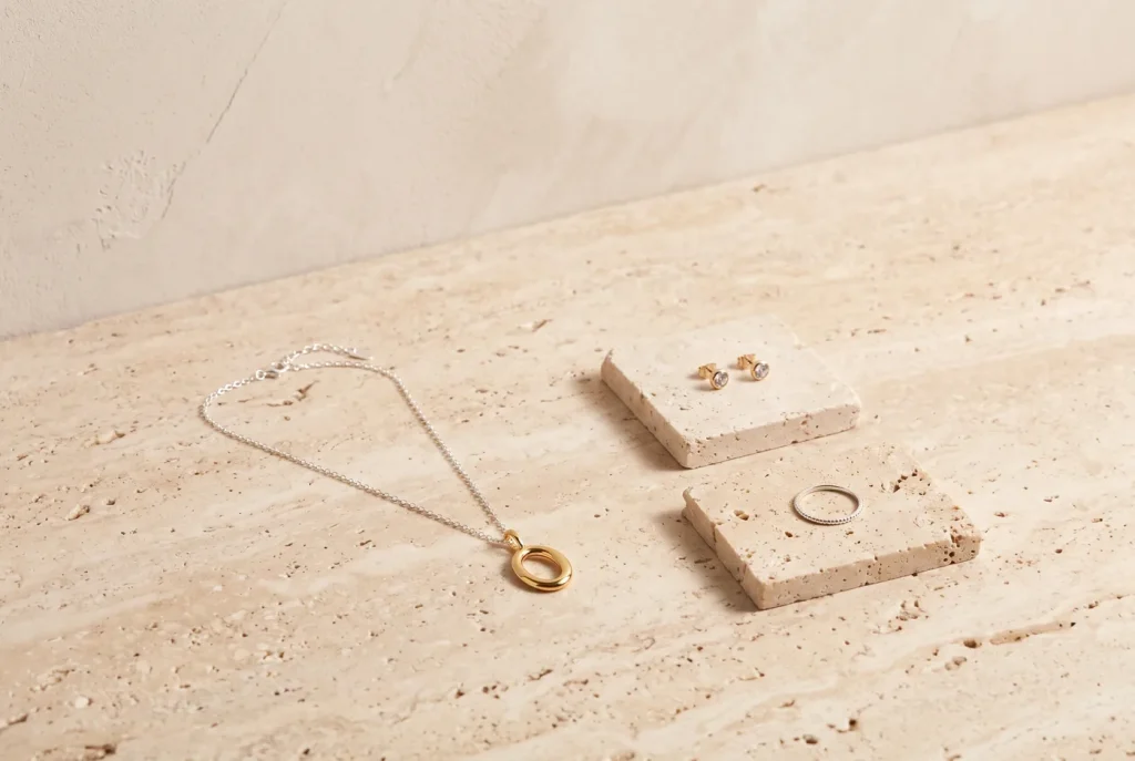 A linen-bound notebook, brass pen, folded fabric swatches in muted tones, and a dish of wooden beads on a travertine surface