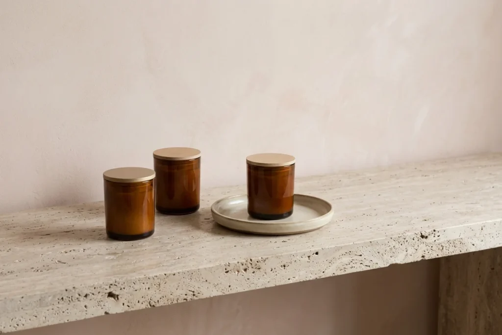 Three amber glass candle vessels and a ceramic tray in an edited grouping on a travertine bench with open space beside them