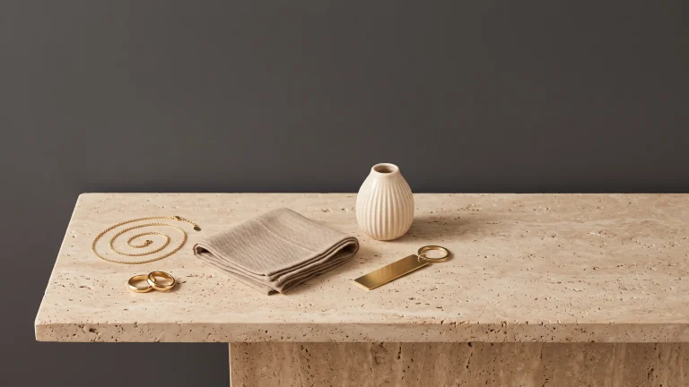 A coiled gold chain necklace, two gold rings, a folded taupe knit scarf, a fluted cream ceramic vase and a brass keychain arranged on a travertine table against a charcoal wall