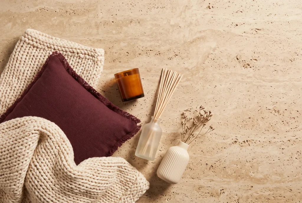 A cream chunky knit throw, a burgundy fringed cushion, an amber candle, a reed diffuser and a ribbed cream vase shot overhead on travertine