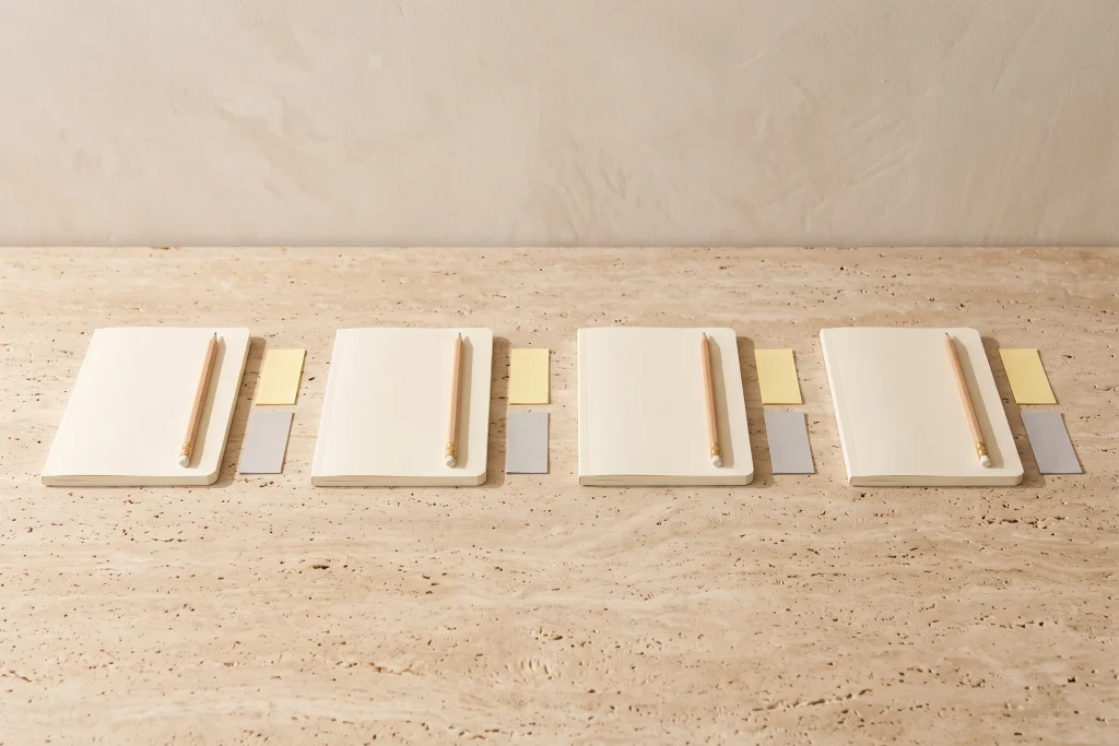 Four matching student gift sets in a row, each with a blank cream notebook, a wooden pencil and small yellow and grey paper notes, on travertine