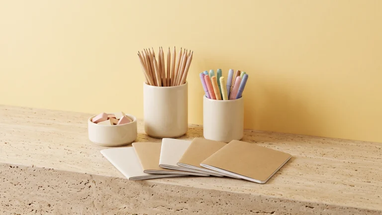 Cream ceramic pots holding pencils, colored markers and erasers with blank kraft notebooks fanned in front, on travertine against a soft yellow wall