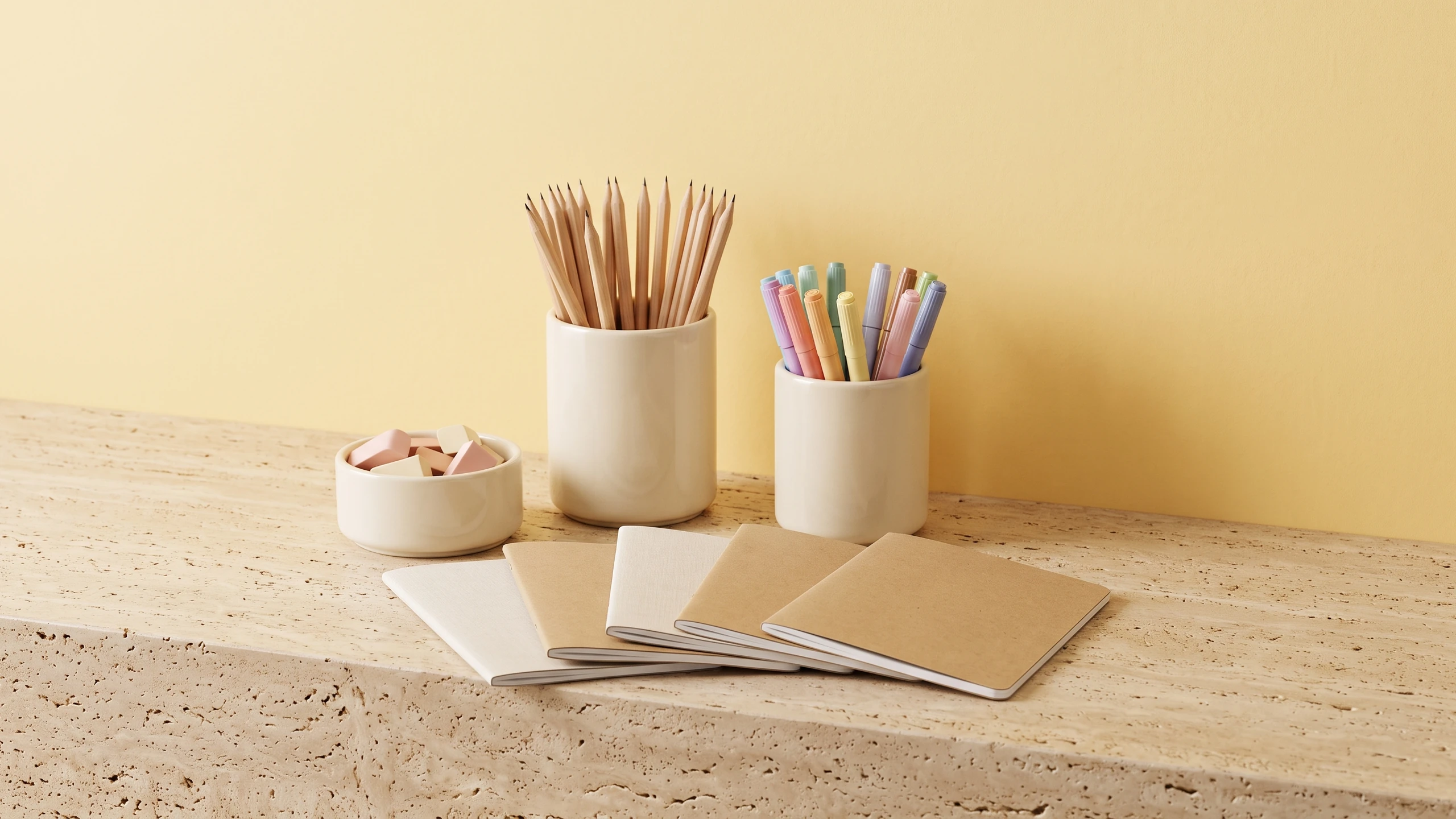 Cream ceramic pots holding pencils, colored markers and erasers with blank kraft notebooks fanned in front, on travertine against a soft yellow wall